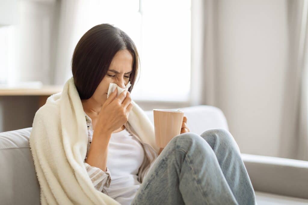 Young woman on sofa, wrapped in blanket, blowing nose and holding mug, due to sinus infection, St. Louis, MO