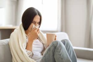 Young woman on sofa, wrapped in blanket, blowing nose and holding mug, due to sinus infection, St. Louis, MO